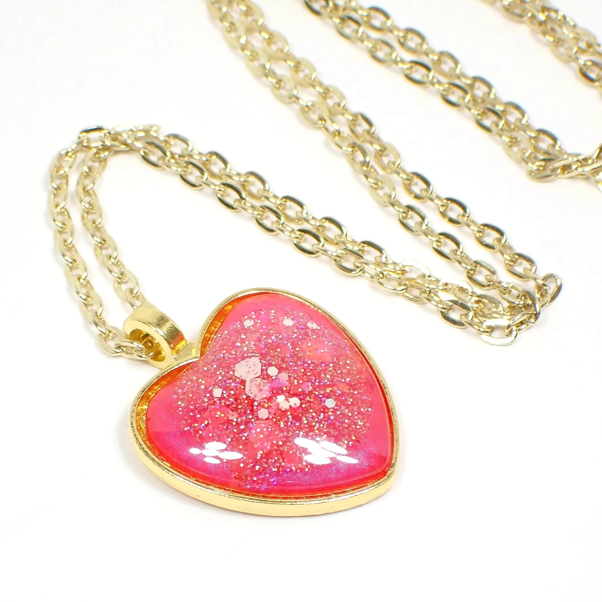 Enlarged view of the gold plated handmade resin heart necklace. The metal is gold in color. The heart has bright pearly pink resin with chunky iridescent glitter. 