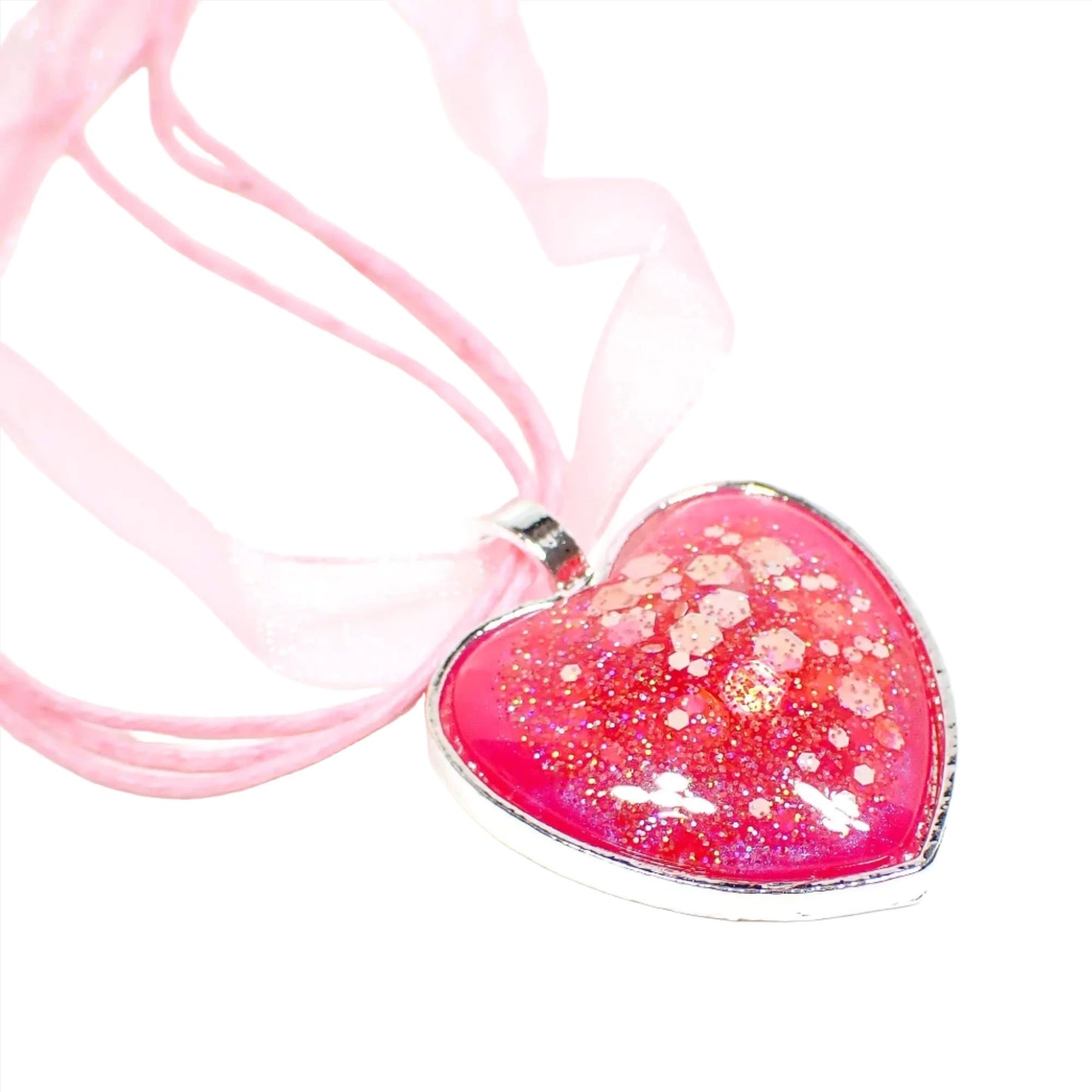 Enlarged view of the silver plated handmade resin heart pendant necklace. The necklace has three strands of light pink faux leather cord and a strand of organza ribbon. The heart has a silver color setting with bright pearly pink resin and chunky iridescent glitter.