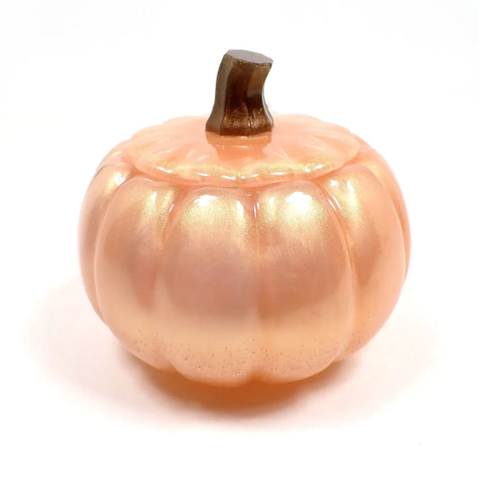 Side view of the handmade resin pumpkin trinket box jar. The pumpkin is a light pastel pearly peach and pink color with a golden sheen. the stem is pearly brown in color. You can see where the lid sets onto the rest of the pumpkin.
