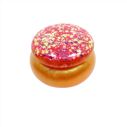 Angled view of the small handmade resin trinket jar. It has a rounded shape. The bottom has pearly orange and gold color resin. The lid has the same color resins and has a lot of chunky iridescent glitter in yellow and pink for lots of sparkle.