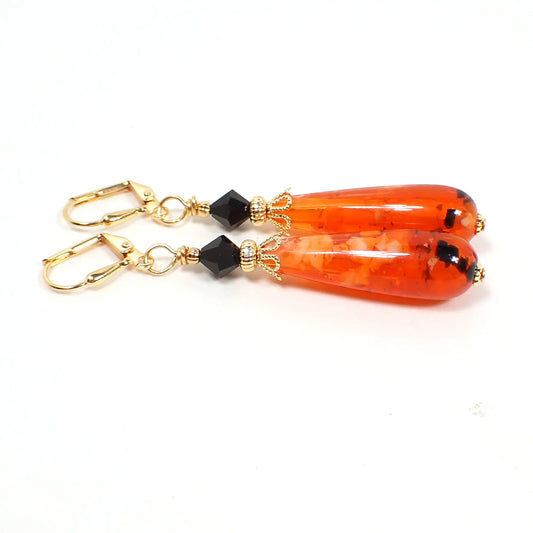Side view of the handmade Halloween earrings with vintage confetti lucite beads. The metal is gold plated in color. The top beads are faceted black glass. The bottom lucite beads are teardrop shaped and are primarily orange in color with small chunks of white and black. Each beads is different from the other in pattern.