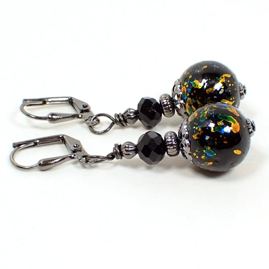 Side view of the handmade metallic splash beaded earrings. The metal is gunmetal gray in color. There are faceted black glass beads on the top. The bottom beads are black round glass and have a splash like design with metallic gold, silver, green, and blue colors.