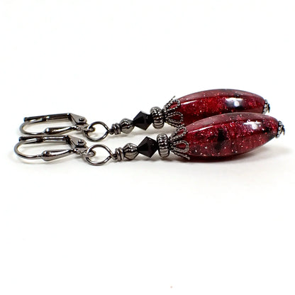 Side view of the handmade oval drop earrings with vintage lucite beads. The top beads are black faceted glass bicone beads. The bottom beads are longer style faceted oval in shape. The lucite has marbled shades of red and pink with black and there are tiny specks of glitter giving them a galaxy like appearance. The metal is gunmetal gray in color.