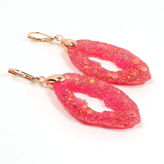 Angled view of the handmade resin geode earrings. They are shaped like a druzy slice with an open middle area. The resin is bright pink with iridescent chunky glitter. The metal is rose gold plated In color.