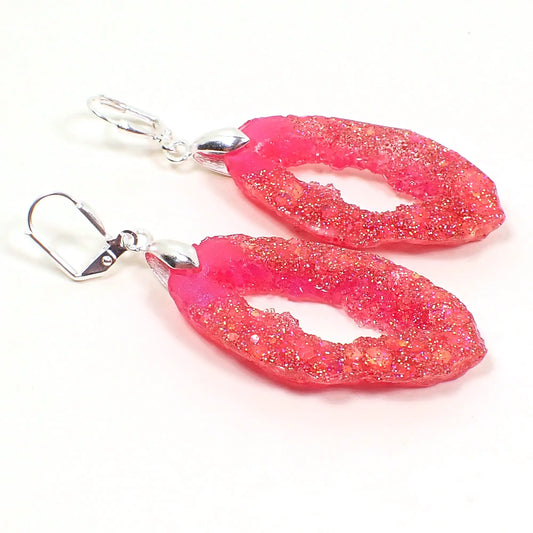 Angled view of the handmade resin geode earrings. They are shaped like a druzy slice with an open middle area. The resin is bright pink with iridescent chunky glitter. The metal is silver plated In color.