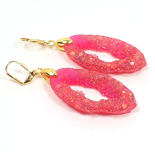 Angled view of the handmade resin geode earrings. They are shaped like a druzy slice with an open middle area. The resin is bright pink with iridescent chunky glitter. The metal is gold plated In color.