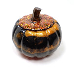 Side view of the handmade resin pumpkin trinket box. The lid has a pearly brown stem and the rest of the lid is black with iridescent chunky glitter that flashes shades of yellow, orange, and red. The pumpkin part has marbled areas of orange and black and a small area of pearly red at the bottom.