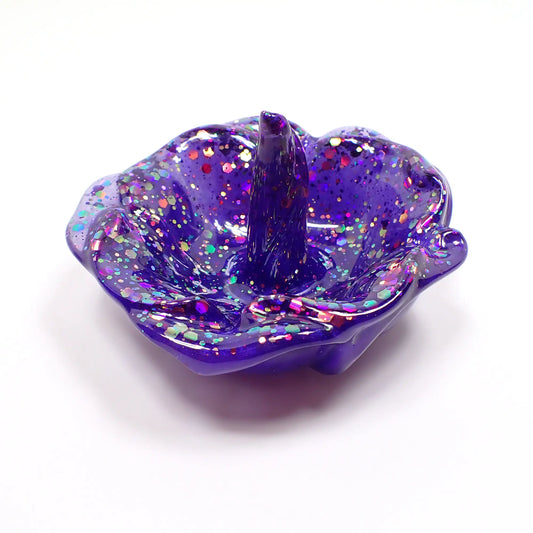Angled side view of the handmade ring dish holder. It is shaped like an open flower with petals around the edge and a single curled petal in the middle that stands up for rings to sit on. The resin is a pearly blue color with flashes of purple depending on how the light hits it. There is chunky iridescent pink, purple, and blue glitter throughout.