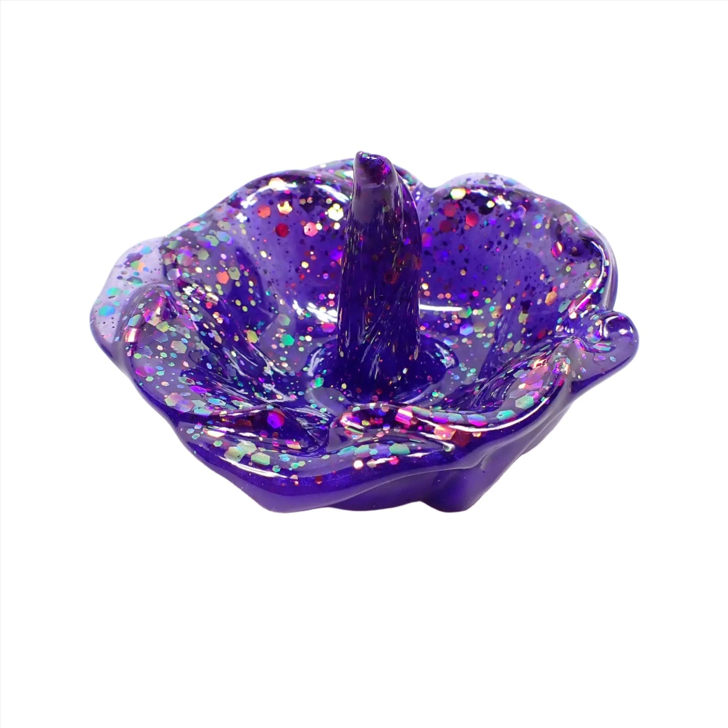 Angled side view of the handmade ring dish holder. It is shaped like an open flower with petals around the edge and a single curled petal in the middle that stands up for rings to sit on. The resin is a pearly blue color with flashes of purple depending on how the light hits it. There is chunky iridescent pink, purple, and blue glitter throughout.