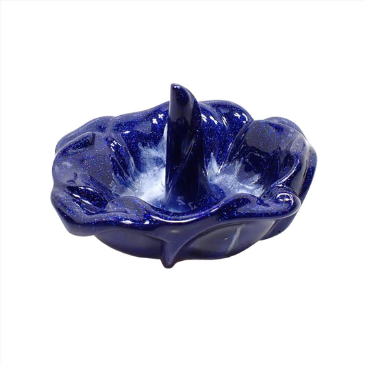 Angled side view of the handmade resin ring dish holder. It is shaped like an open flower with petals around the edge and a single curled petal in the middle that's standing straight up to put rings on. It is mostly pearly dark blue resin with some solid white marbled in around the bottom area. There are tiny flecks of bright blue glitter throughout.