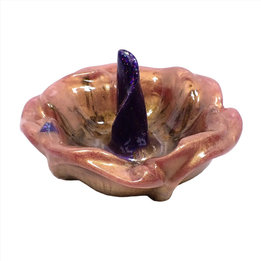 Angled side view of the handmade resin ring dish. It is shaped like an open flower with petals around the edge. There is a area in the middle shaped like a curled petal that is standing straight up for rings to slide over and sit upon. The outer petals are marbled with shades of pink, light purple, and brown. The inside petal has shades of vivid purple and blue with iridescent glitter.