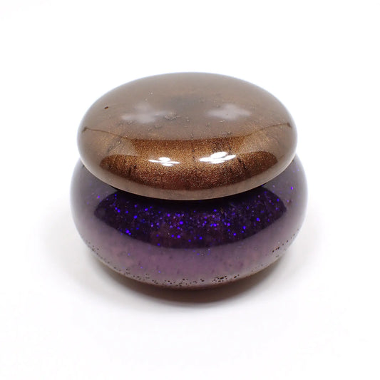 Side view of the small handmade resin trinket box. It has a short rounded jar like shape. The lid is pearly brown in color. The bottom has mostly pearly purple resin with tiny flecks of blue glitter and small area of pearly brown at the very bottom.