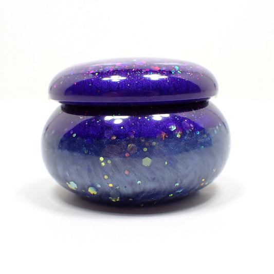 Side view of the small handmade resin trinket box. It is short round jar shaped with a lid. The top part of the piece has vivid pearly blue resin with chunky iridescent purple glitter and the bottom part has a lighter pearly blue resin with the same glitter. 