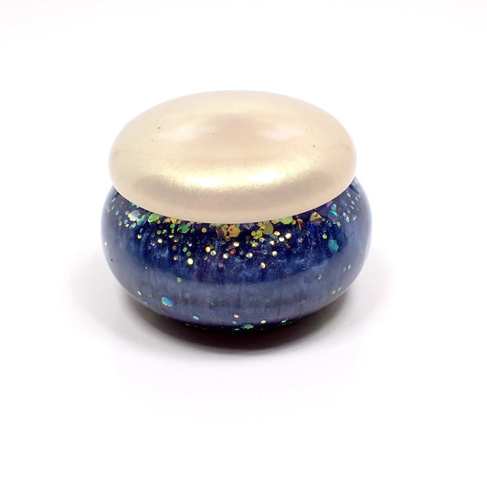Side view of the handmade small trinket box with lid. It is round jar shaped. The bottom part is pearly blue in color with iridescent chunky resin. The lid is off white with a golden sheen.