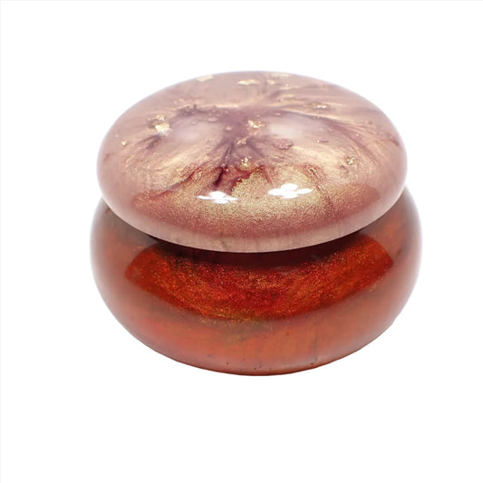 Side view of the small handmade resin trinket box. It has a short rounded jar shape. The bottom part has marbled shades of red, orange, and yellow resin. The top has shades of purple and gold with flakes of metallic gold.