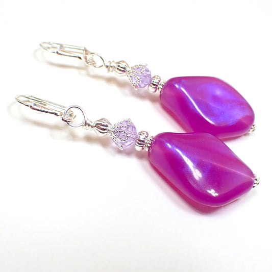 Angled view of the handmade iridescent color shift lucite drop earrings. The metal is silver plated in color. There is a light purple faceted glass crystal bead at the top. The bottom beads are vintage lucite and shaped like angled teardrops. They are a bright purple in color with areas of darker purple as you move around in the light.