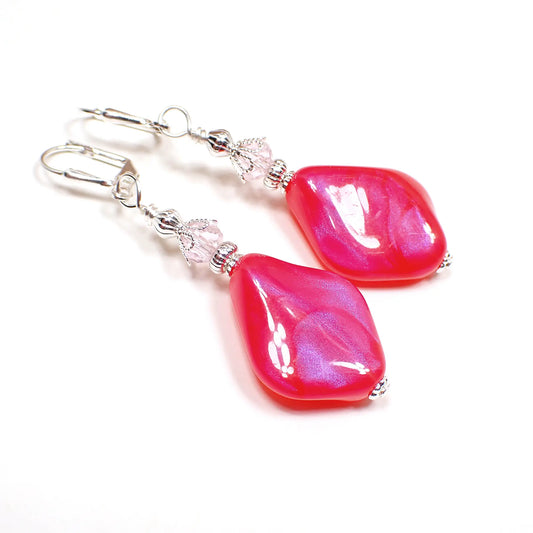 Angled view of the handmade drop earrings with vintage lucite beads. The metal is silver plated in color. There are faceted light pink glass crystal beads at the top. The bottom lucite beads are angled teardrop shaped and are bright pink in color with flashes of purple as the light hits it.