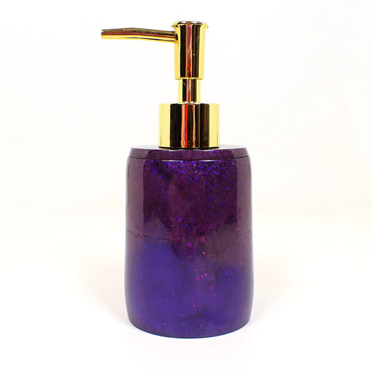Side view of the handmade resin and glitter soap dispenser. It is oval shaped with a gold tone metallic plastic pump style top.There is dark pearly purple resin at the top with tiny flecks of blue glitter. The rest of the dispenser is pearly with a few bits of chunky iridescent glitter.
