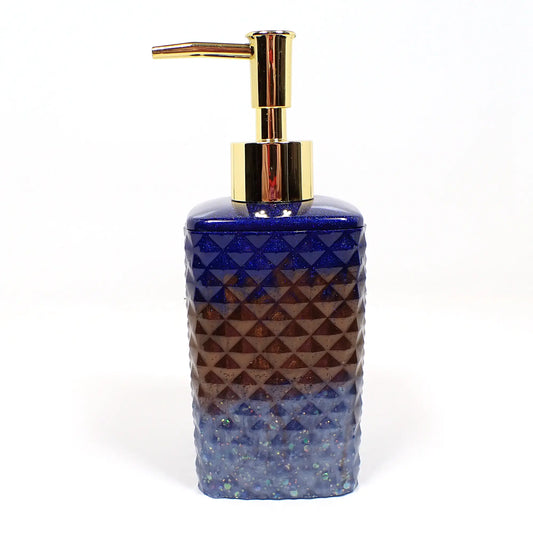Side view of the handmade resin soap dispenser. It has a tall faceted square shape and has a gold color pump on top in this photo. The top part of the dispenser is dark blue with tiny blue glitter, the middle is dark brown, and the bottom is light blue with chunky iridescent glitter.