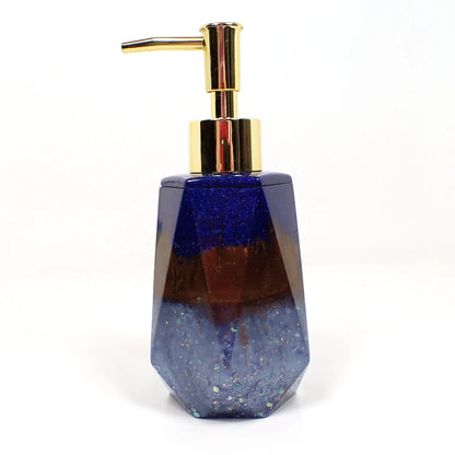 Side view of the handmade resin soap dispenser. In this photo it has a gold tone color pump on it. The top part of the dispenser has pearly dark blue resin with tiny flecks of blue glitter. The middle of the dispenser has pearly brown resin and the bottom has pearly light blue resin with chunky iridescent glitter. It has a faceted shape that is narrower at the top and flared at the bottom.