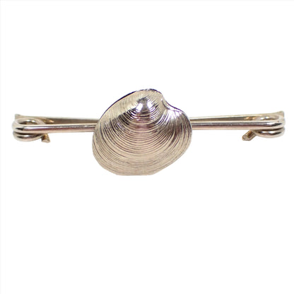 Front view of the retro vintage collar clip. The metal is silver tone in color and the bar has coil ends. There is a seashell shape in the middle of the bar.