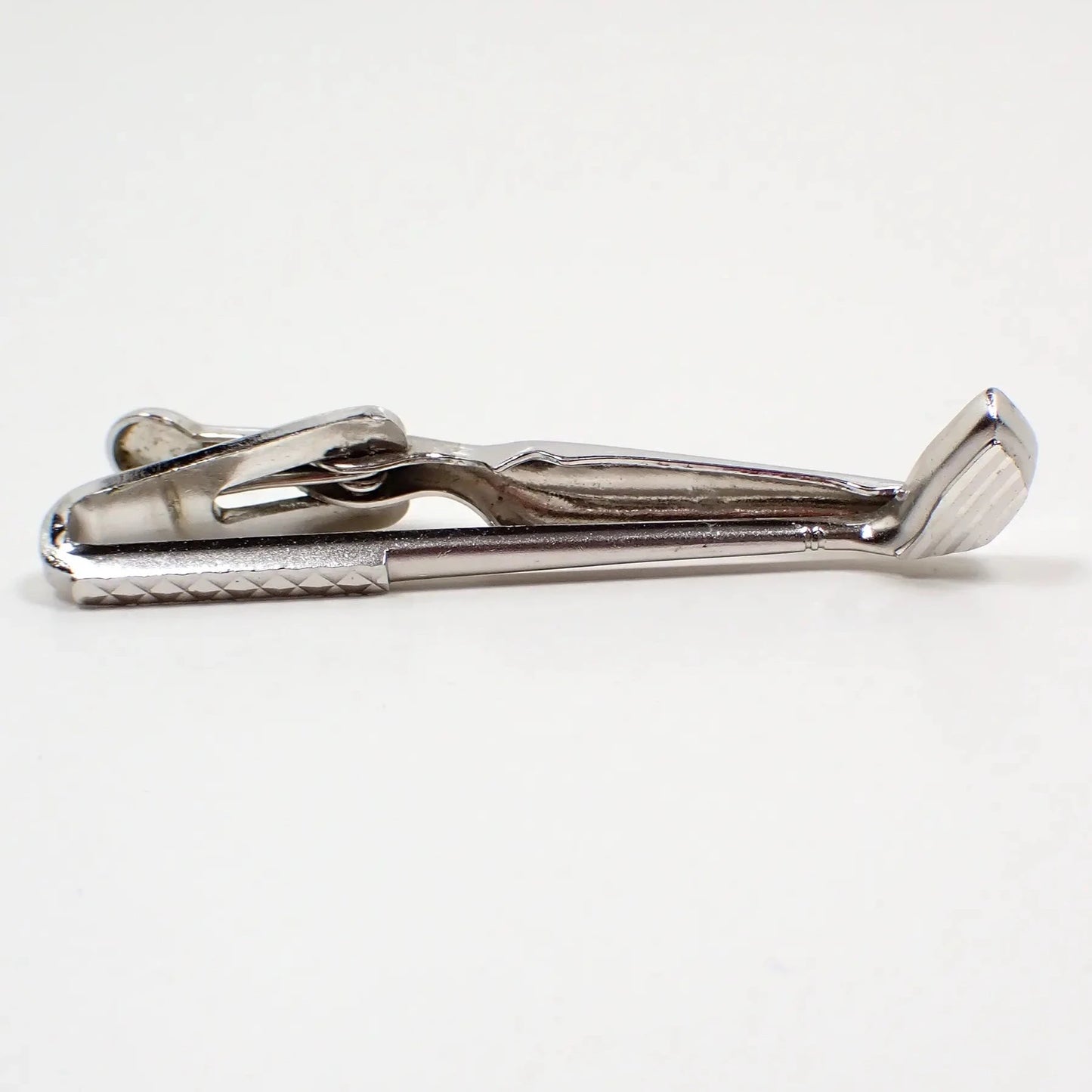 Front view of the retro vintage Swank tie clip. The metal is silver tone in color. It's shaped like a golf club. The handle and bottom part of the club have a cut etched design that gives it some flash as you move around in the light.