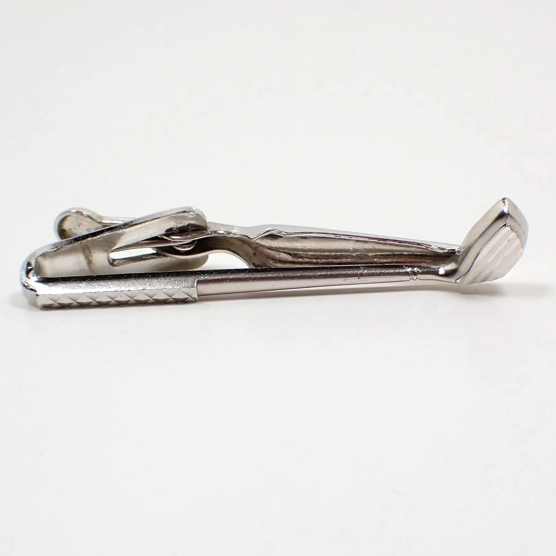 Front view of the retro vintage Swank tie clip. The metal is silver tone in color. It's shaped like a golf club. The handle and bottom part of the club have a cut etched design that gives it some flash as you move around in the light.