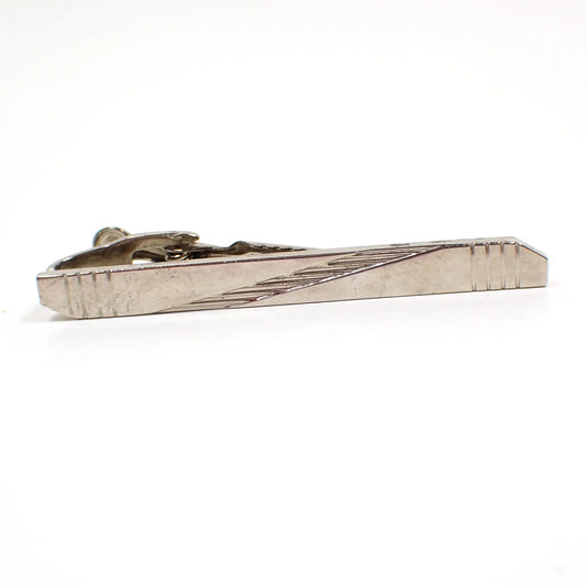 Front view of the retro vintage tie clip. The metal is silver tone in color. It has a long bar like shape with angled ends. There are four engraved stripes on each end and the middle has a textured diagonal stripe.