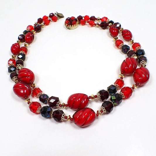 Top view of the West Germany Mid Century vintage multi strand necklace. There are two beaded strands with faceted and oval plastic beads in shades of dark and bright red. There is a round box clasp at the end.