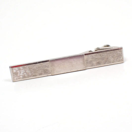 Angled view of the Krementz retro vintage tie clip. The metal is silver tone in color. It's shaped like a rectangle bar with a textured matte tiny line design. The middle has a plain shiny rectangle. A thin scratch can be seen in the middle area under magnification.