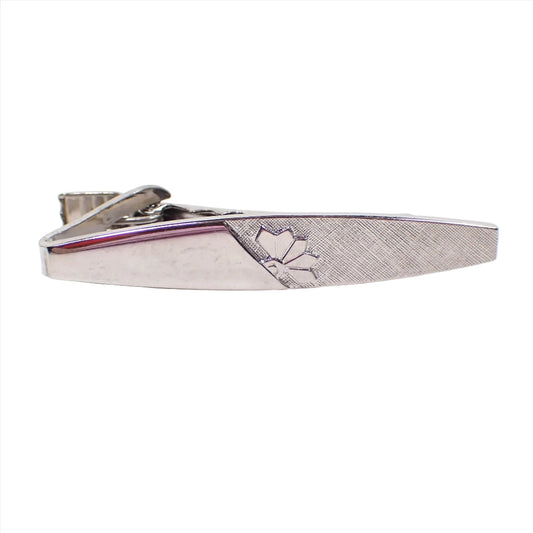 Front view of the retro vintage tie clip clasp. It is marquis shaped with squared off ends. The metal is silver tone in color. The left side has shiny metal and the right side has textured matte metal with half a flower design on it. The two types of metal come together in the middle at a diagonal edge.