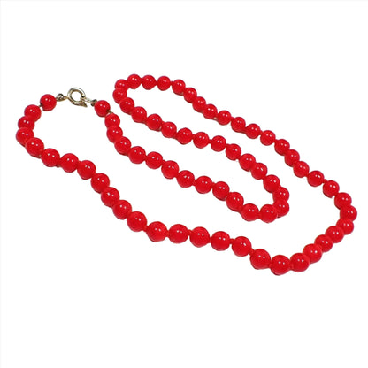 Angled view of the retro vintage glass beaded necklace. The necklace is beaded with small round glass beads in red that are hand knotted in between each bead. There is a gold tone color spring ring clasp at the end.
