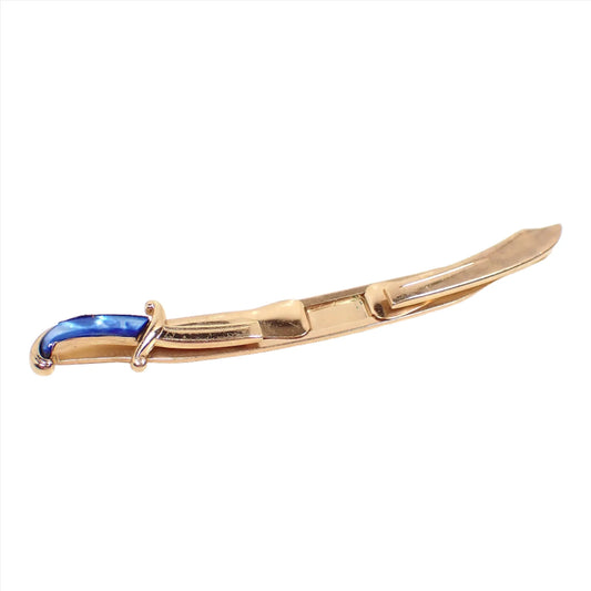 Angled view of the Swank Mid Century vintage sword tie bar. It's shaped like a scimitar with gold tone color metal. The hand has blue dyed mother of pearl shell. There is an open area on each side to slide the tie into.