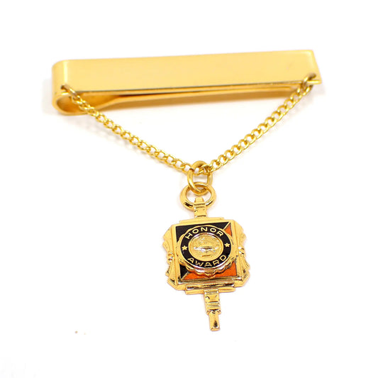 Front view of the vintage slide on tie chain with charm. The metal is gold tone in color. The chain is curb link. The charm hanging on the chain says Honor Award and has black and orange enamel on it.