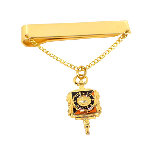 Front view of the vintage slide on tie chain with charm. The metal is gold tone in color. The chain is curb link. The charm hanging on the chain says Honor Award and has black and orange enamel on it.
