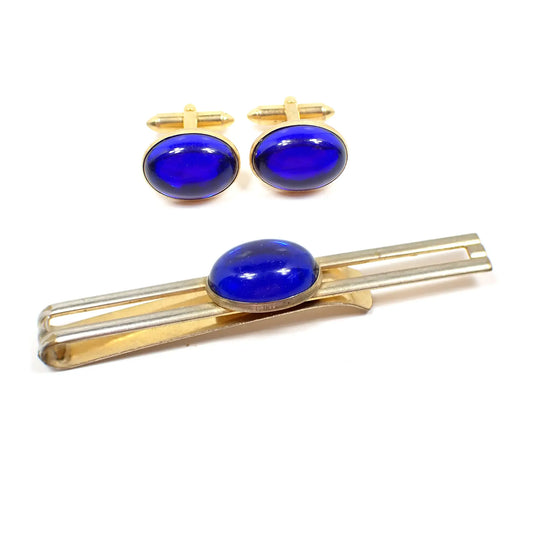 View of the Hadley Mid Century vintage men's jewelry set. The metal is gold tone in color. There is a slide on tie bar and cufflinks. The tie bar has an open bar style with oval lucite cab in the middle in bright blue. The cufflinks are also oval with blue lucite cabs. 
