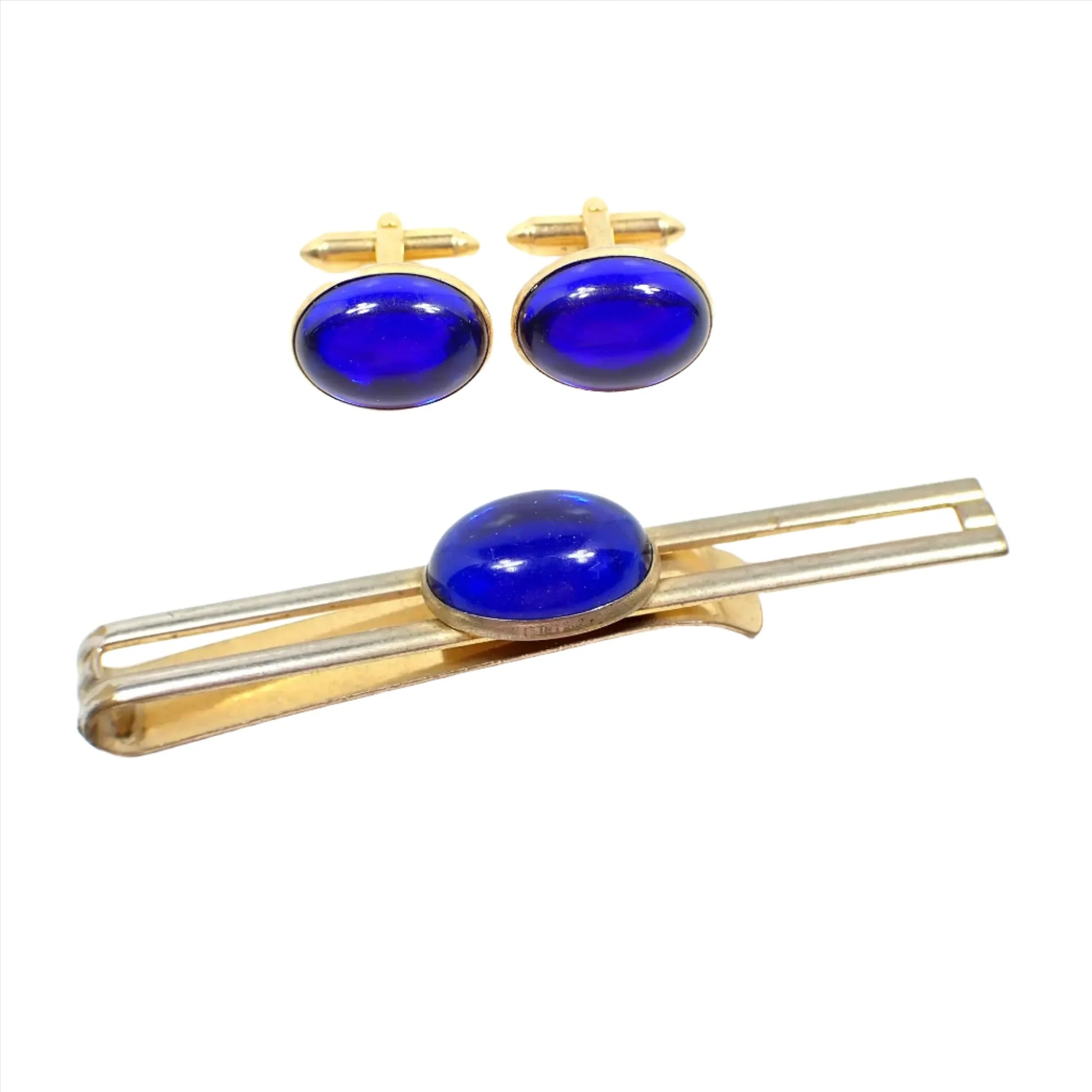 View of the Hadley Mid Century vintage men's jewelry set. The metal is gold tone in color. There is a slide on tie bar and cufflinks. The tie bar has an open bar style with oval lucite cab in the middle in bright blue. The cufflinks are also oval with blue lucite cabs. 