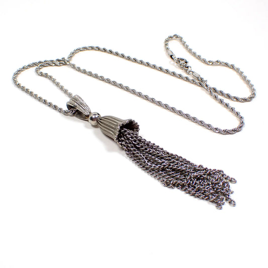 Angled view of the retro vintage tassel pendant necklace. The metal is silver tone in color. The chain is a twisted rope chain with a lobster claw clasp at the end. The pendant is textured and bell shaped with a large bail at the top. From the bottom of the bell shape are several dangling strands of curb chain to form the tassel.