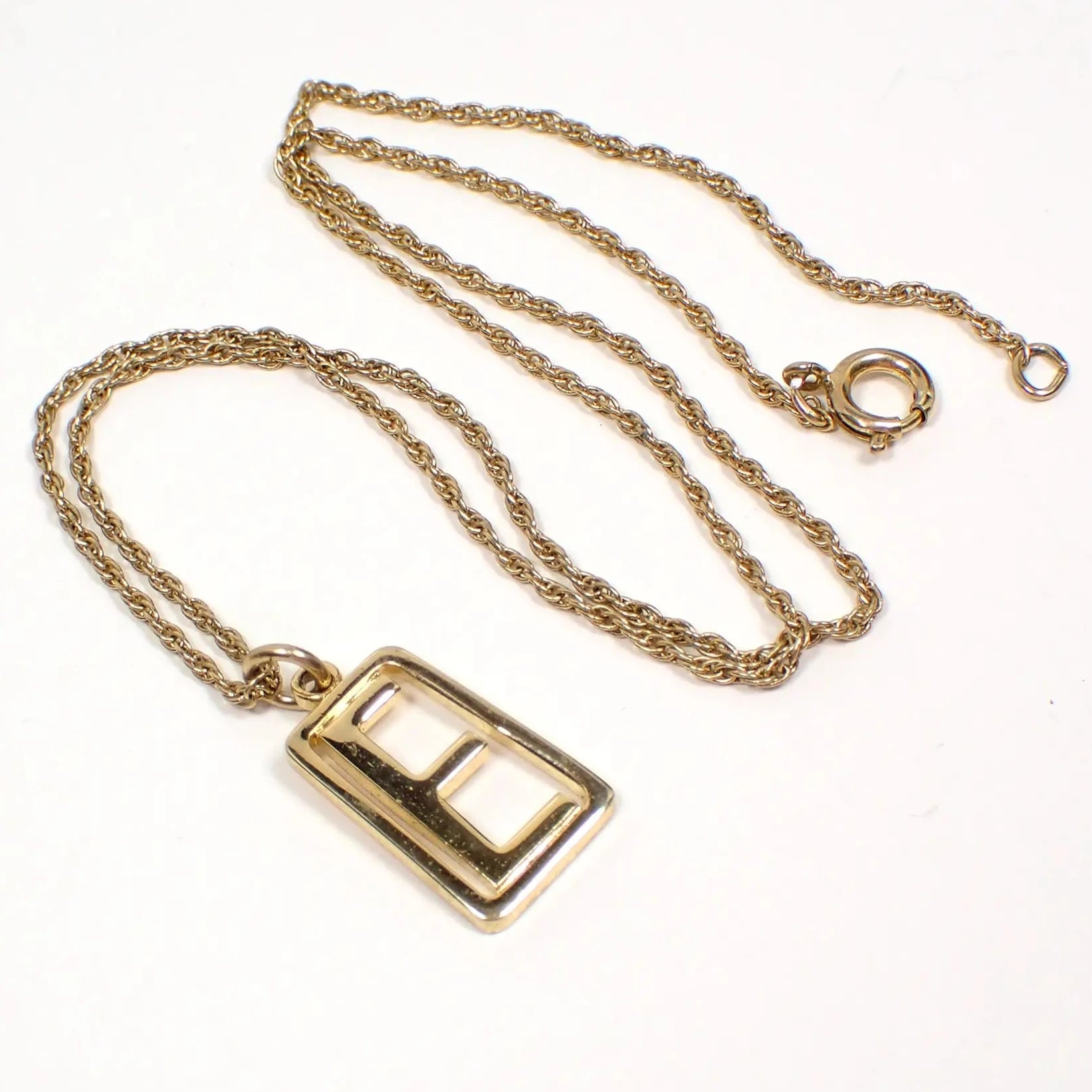 Angled view of the retro vintage initial pendant necklace. The metal is gold tone in color. The rope chain is thinner style with a spring ring clasp on the end. The pendant hangs at the bottom  and is trapezoid shaped with a block style letter E in the middle.