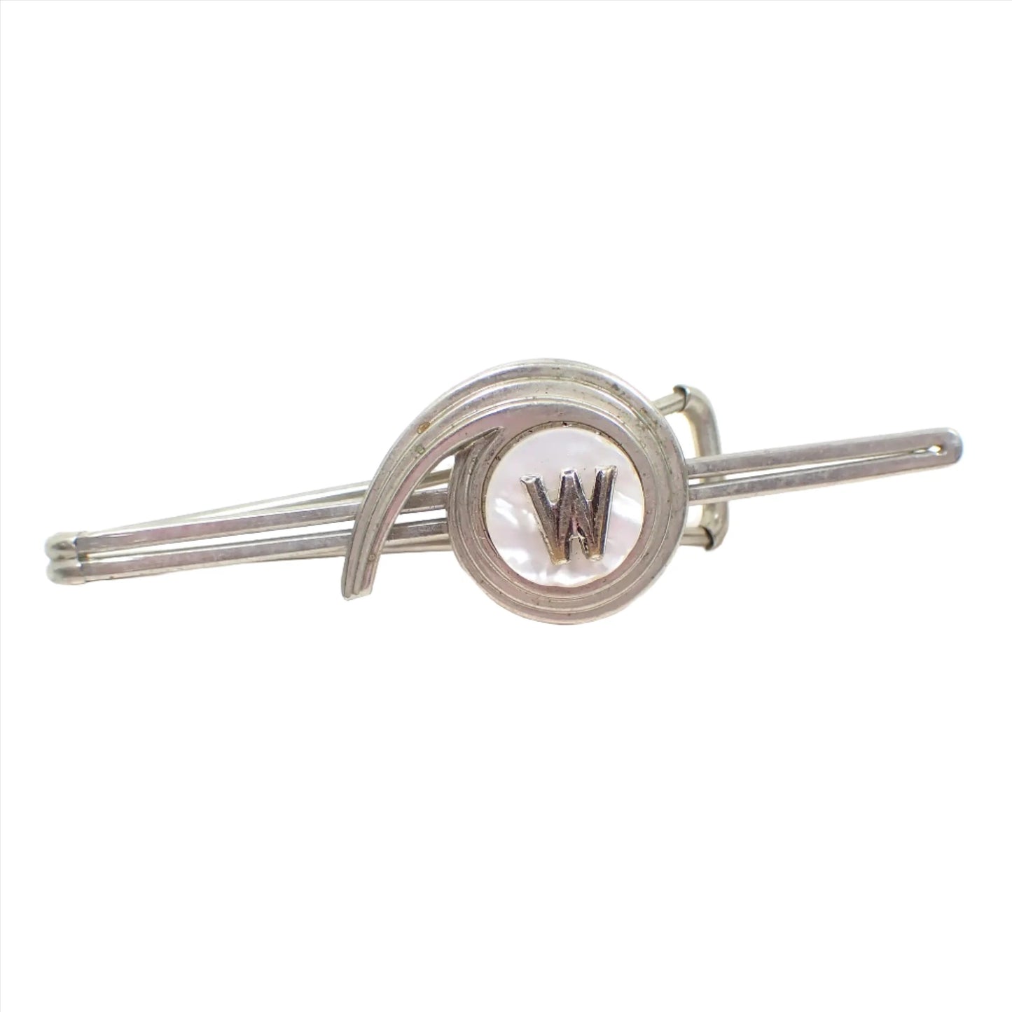 Front view of the Art Deco tie bar. The metal is silver tone in color. It has an open long design with a round area in the middle with the metal design arcing over it and then tapering at the end. The center has a pearly white mother of pearl shell cab and there is a silver tone initial letter W in the very center.