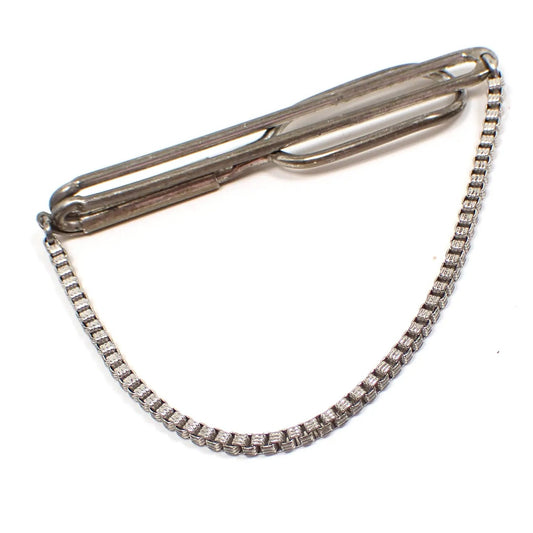 Angled view of the Swank 1930's slide on tie bar chain. The sterling silver has darkened with age to a gray color. The bar has an open long oval design. There is a fancy square link chain hanging on the bottom.