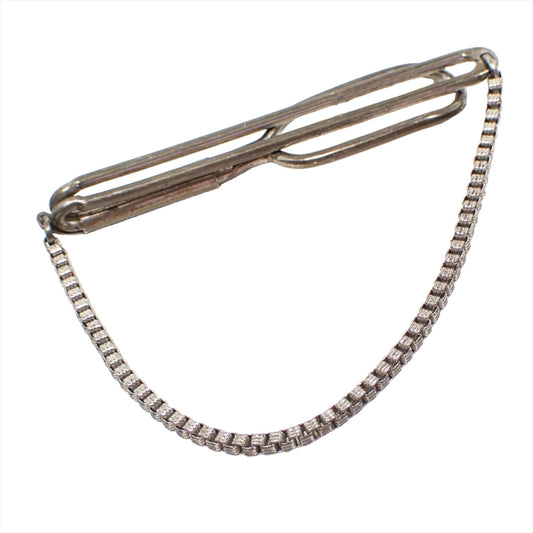 Angled view of the Swank 1930's slide on tie bar chain. The sterling silver has darkened with age to a gray color. The bar has an open long oval design. There is a fancy square link chain hanging on the bottom.
