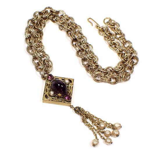 Top view of the Mid Century vintage pendant tassel necklace. The chain is double link cable with a textured pattern and hook clasp at the end. There is a filigree diamond shaped pendant with two purple rhinestones, two faux pearls, and an oval fancy glass cab that is purple with speckles on the inside. There is a chain tassel with faux pearls on the ends at the bottom. The metal is antiqued gold in color.