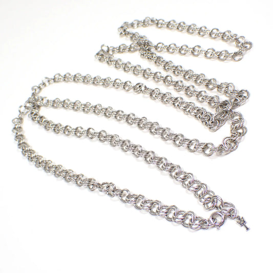 Angled view of the retro vintage Crown Trifar chain necklace. It is silver tone in color. There are double round rolo shaped links throughout the chain. There is a spring ring clasp at the end with a T with a crown on top symbol hang tag for Crown Trifari.