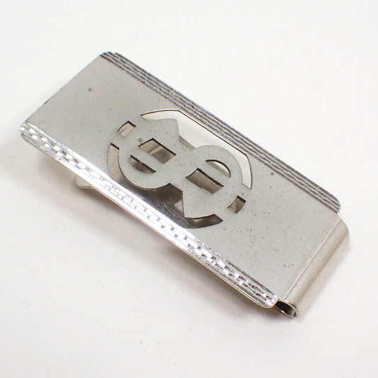 Angled view of the retro vintage money clip. The front is matte silver color with a cut out dollar sign design. The edges have a faceted shiny silver tone appearance.