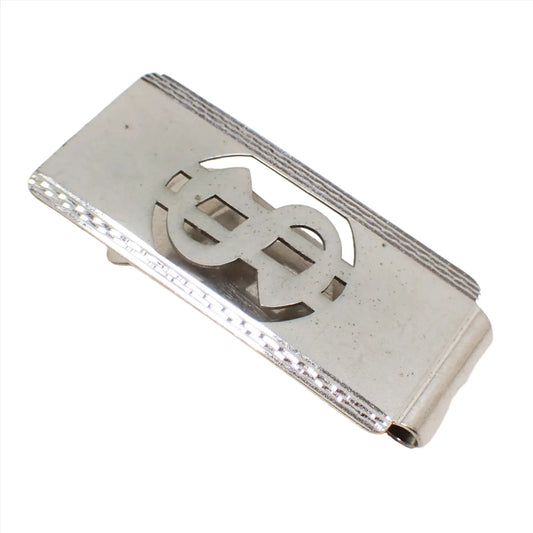 Angled view of the retro vintage money clip. The front is matte silver color with a cut out dollar sign design. The edges have a faceted shiny silver tone appearance.