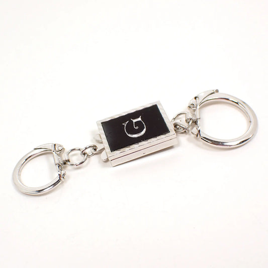 Front view of the Mid Century vintage Swank keychain. It has a rectangle part in the middle with a black plastic front that has an initial letter G in the middle. The rest of the piece is silver tone in color. There is a small key ring on one end with a lock over type clasp and a larger one on the other end. The smaller one has a clip at the end of the ring to remove it from the rest of the piece if desired.