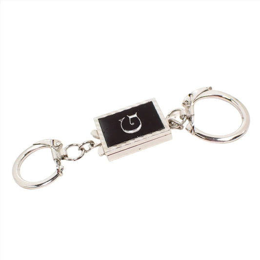 Front view of the Mid Century vintage Swank keychain. It has a rectangle part in the middle with a black plastic front that has an initial letter G in the middle. The rest of the piece is silver tone in color. There is a small key ring on one end with a lock over type clasp and a larger one on the other end. The smaller one has a clip at the end of the ring to remove it from the rest of the piece if desired.