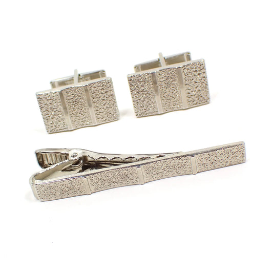 Front view of the Mid Century vintage men's jewelry set. There is a tie clip and cufflinks. The metal is silver tone in color. The cufflinks are rectangle and have a bumpy textured pattern on the front with raised shiny lines. The tie clip is a long rectangle bar shape and has the same pattern.
