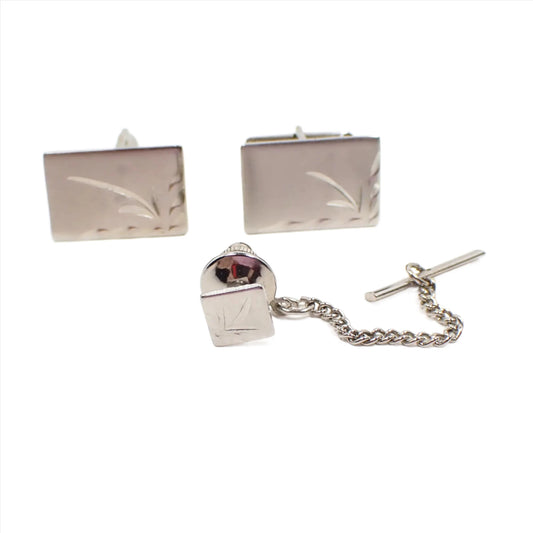 Front view of the Mid Century vintage men's jewelry set. There is a tie tack and a pair of cufflinks. The cufflinks are rectangle shaped and are matte silver color with a shiny etched side and bottom edge and leaf design in the corner. The tie tack has the same design but is square shaped.